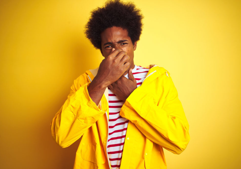 African american man with afro hair wearing rain coat standing over isolated yellow background smelling something stinky and disgusting, intolerable smell, holding breath with fingers on nose. Bad smells concept.