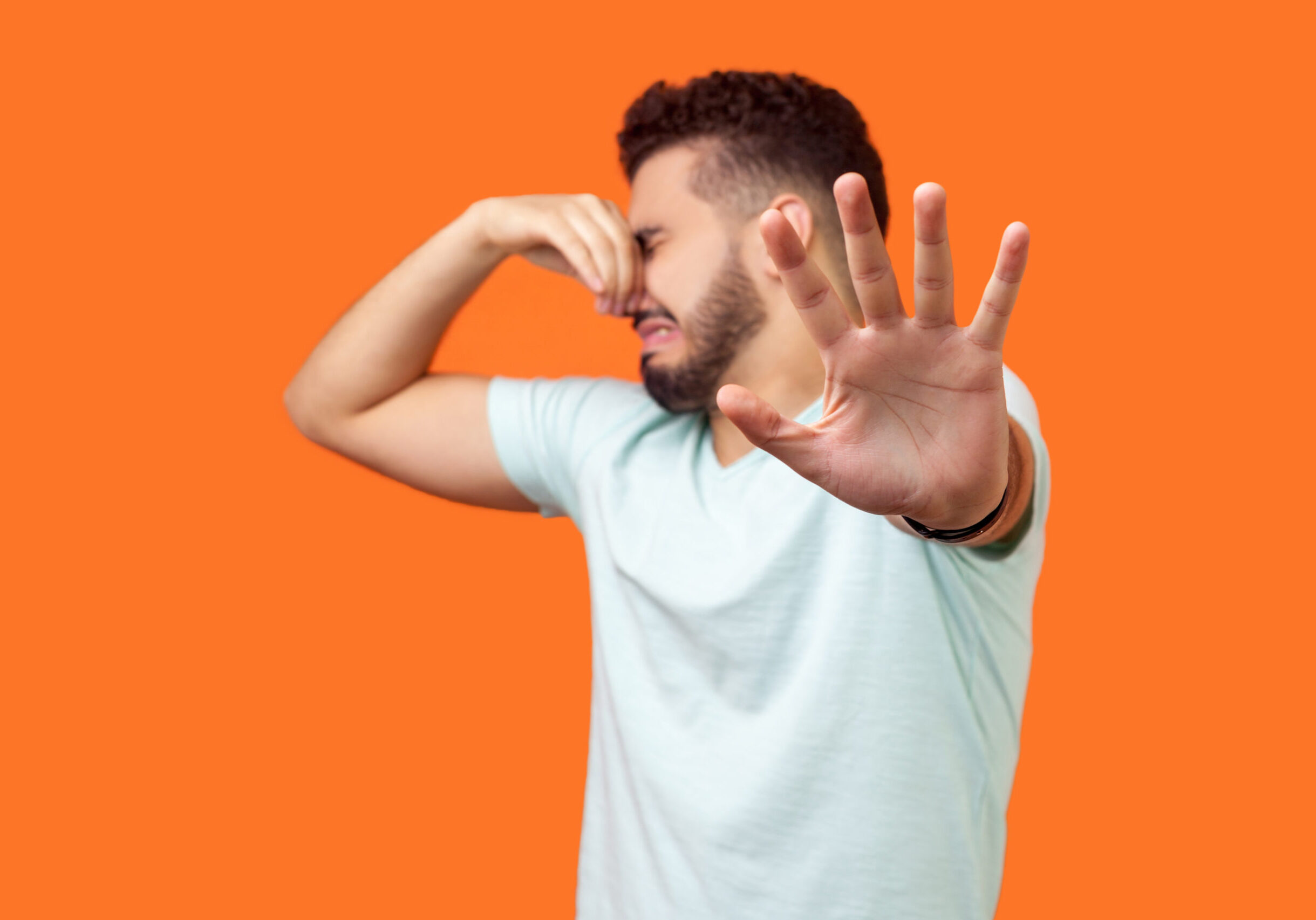 Stinky smell. Side view of confused brunette man with beard in white t-shirt holding breath with fingers on nose, disgusted by bad odor fart, gesturing stop. studio shot isolated on orange background